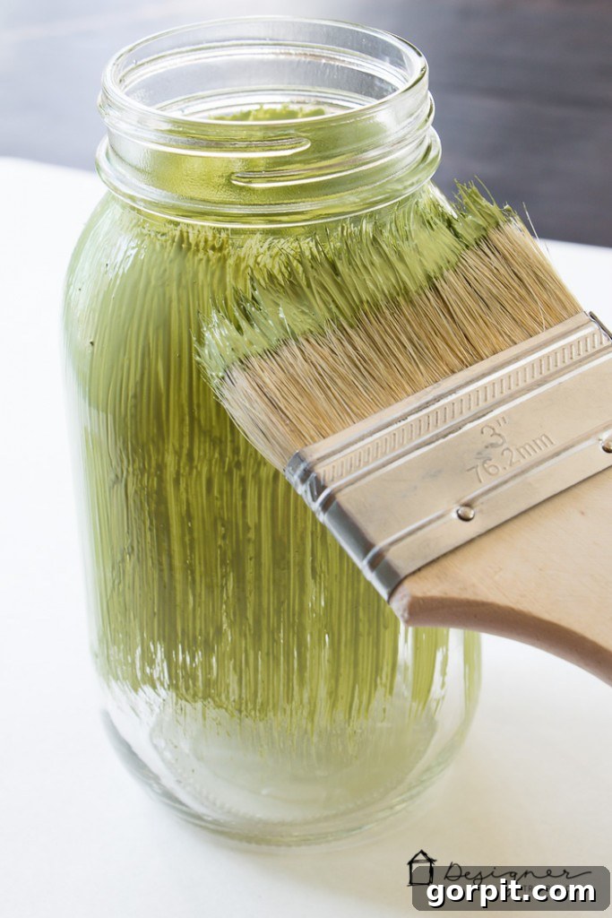 A mason jar with applied vinyl stencil and the first coat of chalk paint, demonstrating the early stages of a personalized gift project.