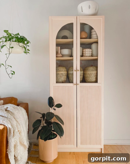 A tall cabinet with arched glass doors next to potted plants, a light leather chair and off-white blanket. This DIY IKEA hack combines Billy bookcases and IKEA Oxberg cabinet doors. 