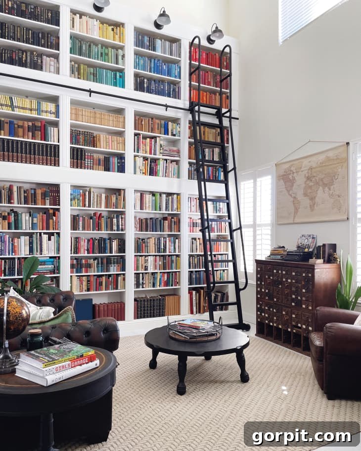 A library in a home with leather chairs, round coffee table, wall map, and an entire backwall of IKEA Billy bookcases converted into a two-story bookshelf, complete with a black iron ladder on wheels. 