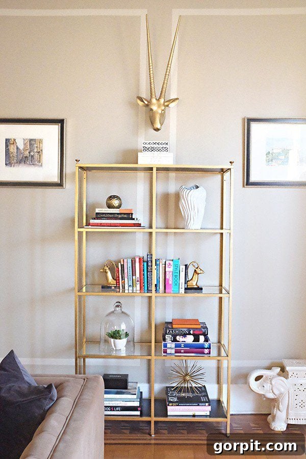 A gold storage rack made from IKEA Vittsjo holds books and other items. A gold horned animal head is above the open shelves, and framed pictures are on either side. 