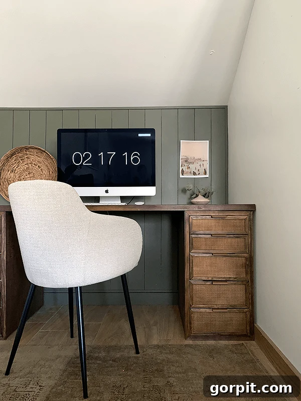 A small desk with a dark stained surface and caned drawers has a computer monitor on top and a modern fabric chair in front. The monitor reads "02 17 16." 