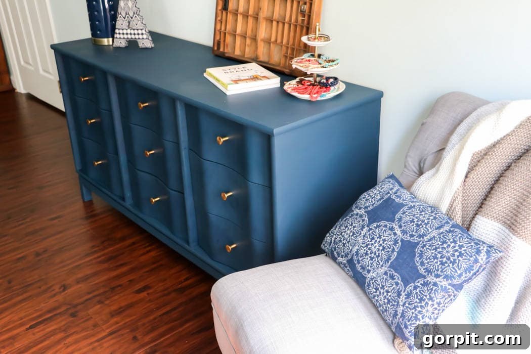 Wood Wonders: Your Guide to Transforming Furniture with Paint 11 Angled view of a navy-blue painted wood dresser with gold knobs, with a lamp. books, a three-tiered tea and cake holder and more on top. A cozy gray armchair with a blue patterned pillow and knit throw is to the right. The simple house outline logo and the website, "DesignerTrapped.com" is in the lower corner. This dresser is an example of how to paint wood furniture.