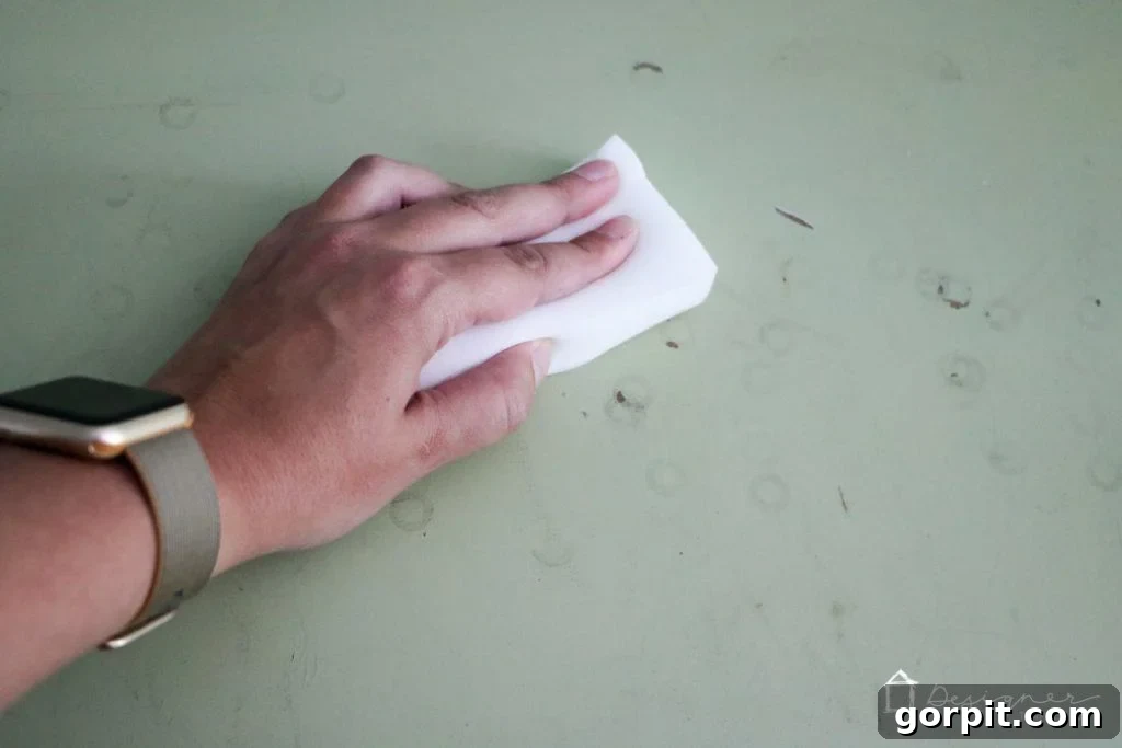 Wood Wonders: Your Guide to Transforming Furniture with Paint 6 A hand with a green and gold watch uses a Mr. Clean Magic Eraser to scrub dirt and stains off an old green dresser top.