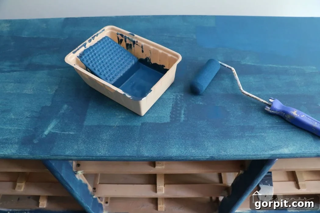 Wood Wonders: Your Guide to Transforming Furniture with Paint 7 An above view of a dresser being painted blue. A paint tray and roller are on the top of the dresser, and the shelves are empty below. The first coat of blue paint is on the top and shelf supports. The corner text reads, "Designer Trapped.com."
