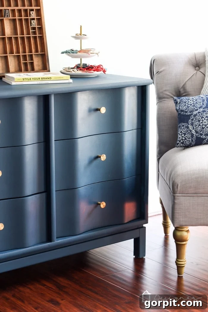 Wood Wonders: Your Guide to Transforming Furniture with Paint 10 Close-up of a navy blue painted wood dresser with gold knobs, styled with a tiered tray of accessories, books, and a vintage letterpress, placed beside a tufted gray armchair. A polished example of how to paint wood furniture for a modern makeover. Text in the lower right corner says, "DesignerTrapped.com" with a drawing of a house as a logo.