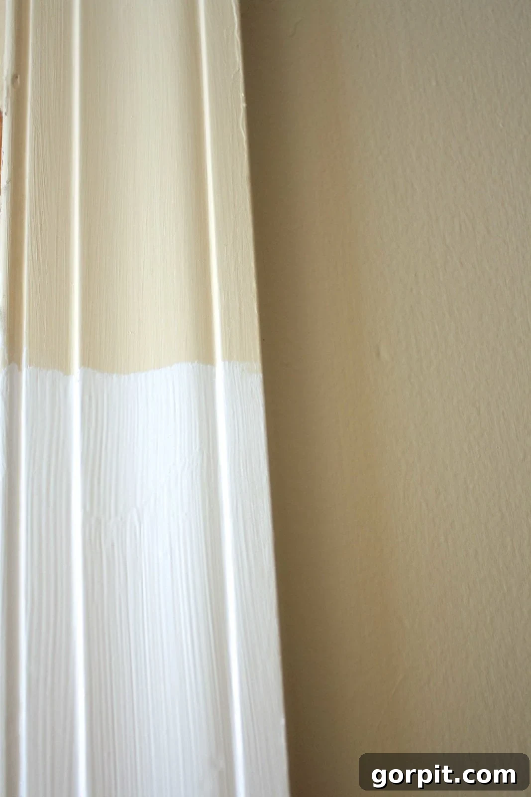 A close-up of door trim, half painted pale beige and the bottom half painted with a new coat of bright white paint. 