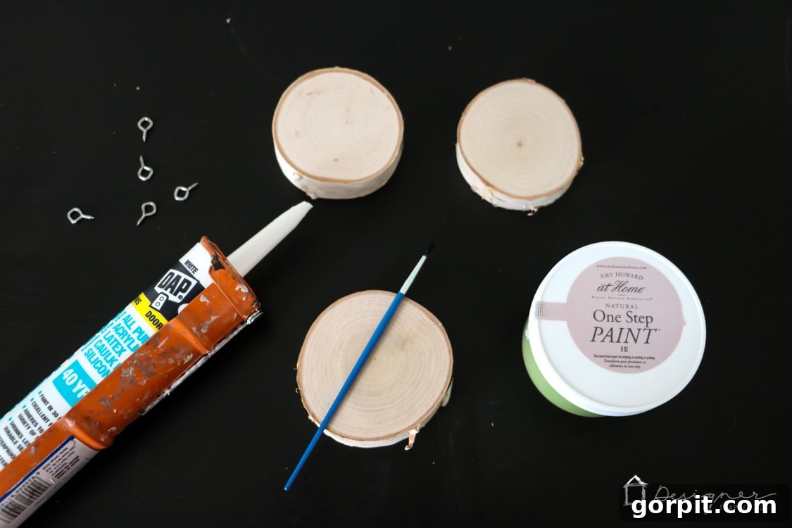 All the crafting supplies neatly arranged: caulk, caulk gun, wood slices, paints, brushes, and screw eyes, ready for making DIY ornaments.
