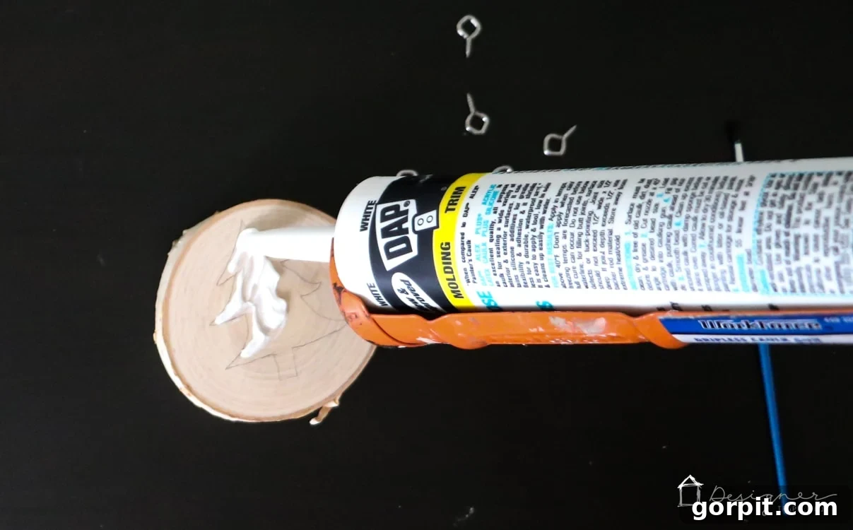A caulk gun being used to apply a white caulk outline over a pencil-sketched Christmas tree on a wooden ornament.