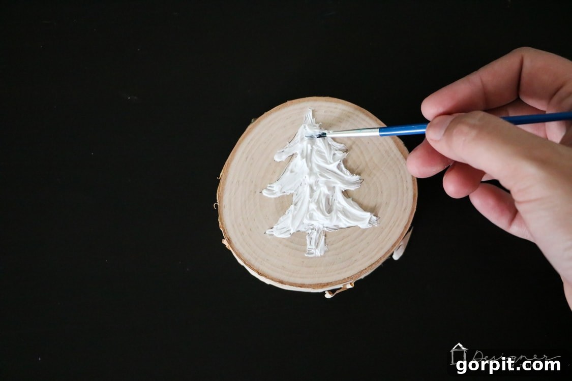 A small paintbrush smoothing and filling the Christmas tree outline with white caulk to create a textured surface on the wood slice.
