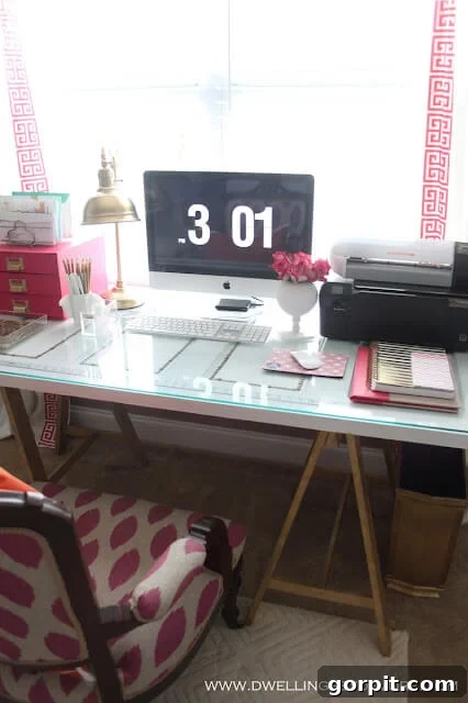 Smart IKEA Desk Upgrades for Less 6 A stylish IKEA desk hack features a repurposed door placed on gold sawhorse legs, topped with glass, along with other office essentials such as a computer monitor, printer, and pink accessories. "www.dwellingsbydevore.com" is printed in the corner.