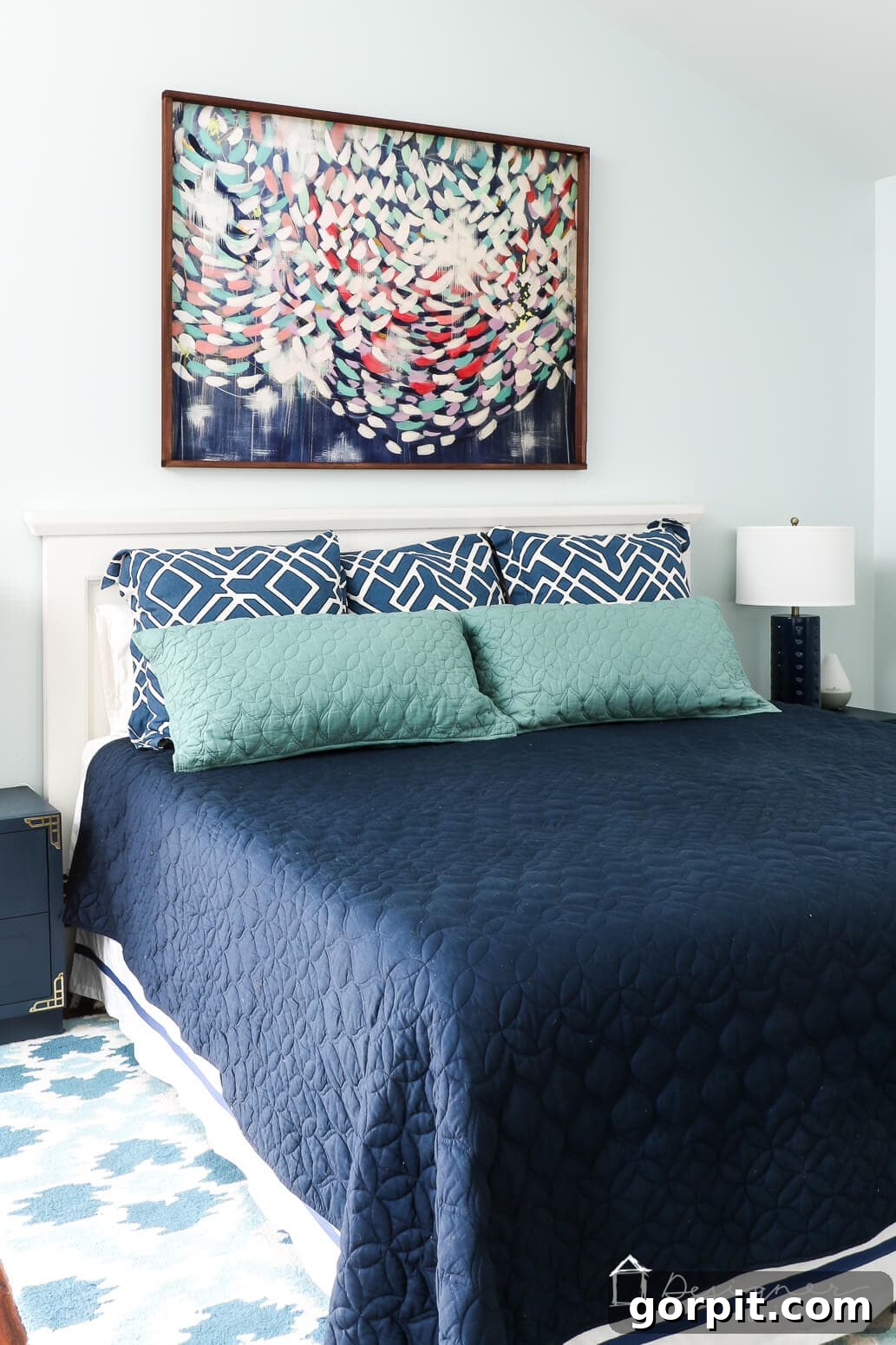 Crafting Stunning Frames for Your Big Art 11 A handmade picture frame encases a large abstract art print above a bed with teal and blue pillows, a blue bedspread, white headboard and a white and blue lamp.