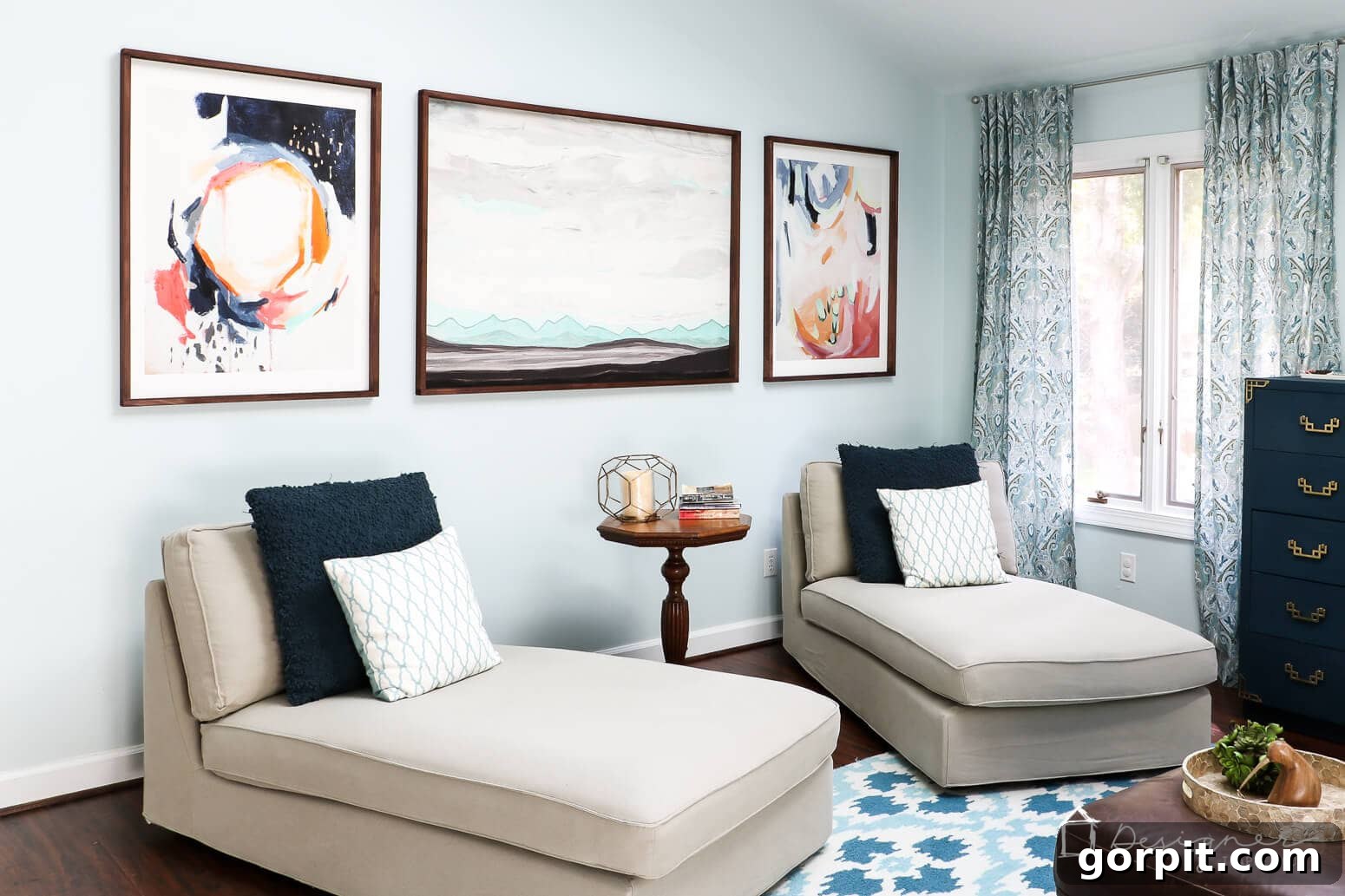 Crafting Stunning Frames for Your Big Art 13 A living room with light blue walls holding three abstract art prints in wooden frames, above two cream-colored chaise loungers with navy and white accent pillows. A round side table separates the chairs and is positioned under the central art piece.
