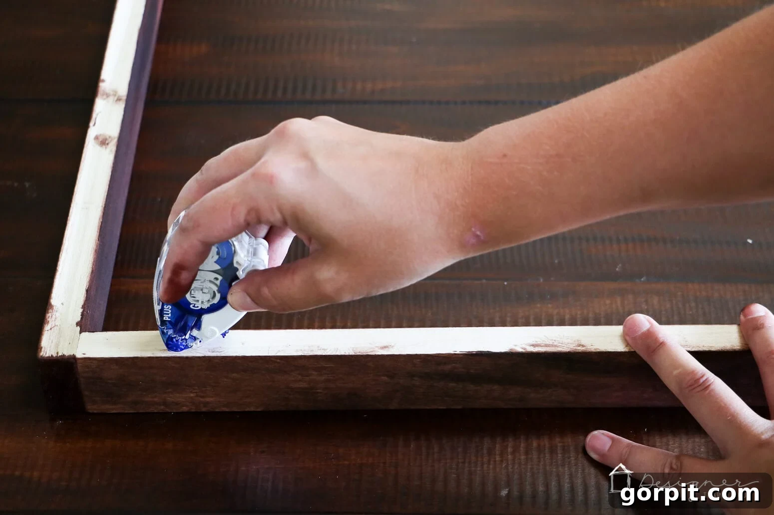Crafting Stunning Frames for Your Big Art 8 Tasha's hand holds a glue tape runner as she applies it to the back of the large art frame resting on a dark wood table. You can learn how to make a picture frame and how to hang an art print in the tutorial.