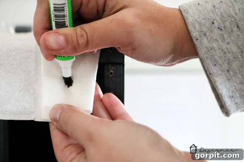 A detailed shot of a woman's hands pressing the tip of a Mohawk 3-in-1 Repair Stick onto a paper towel, illustrating the priming process for easy IKEA furniture repair.