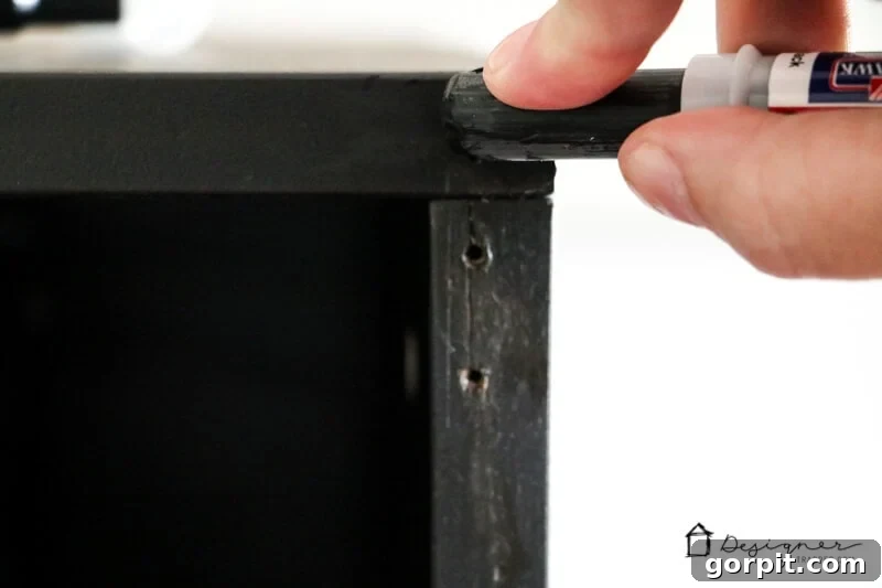 A zoomed-in image showing a hand applying a furniture repair stick to fill nail holes on the back of an IKEA furniture piece. The text "DesignerTrapped.com" with a house logo is visible in the corner.