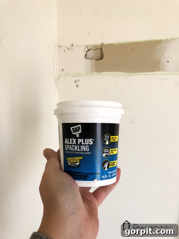 Holding DAP Alex Plus Spackling Paste in front of damaged drywall, ready for repair.