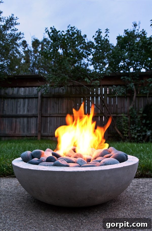 A DIY concrete bowl fire pit sits on gravel in a backyard with a wooden fence, showcasing a modern, minimalist design.