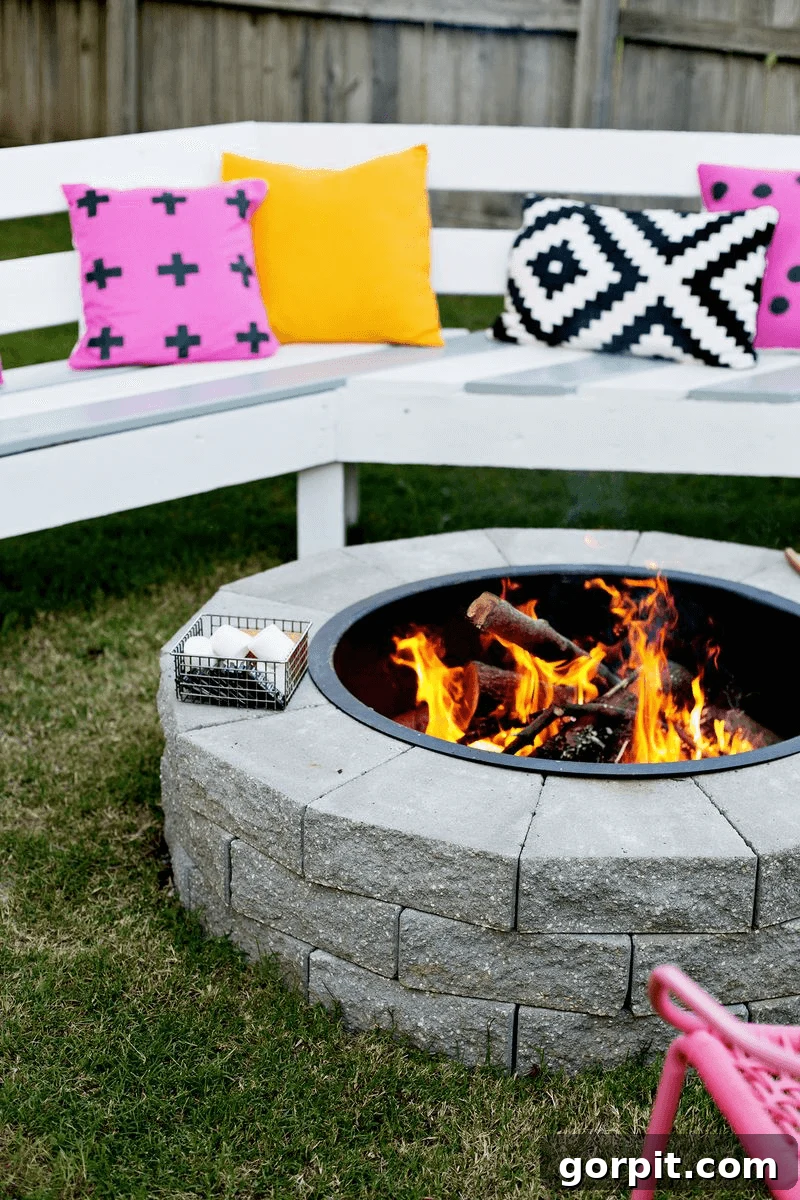The best fire pit ideas are as easy as this DIY fire pit made from landscaping stones, circled by white benches and yellow, pink, and black and white pillows, creating a cozy and colorful gathering spot.
