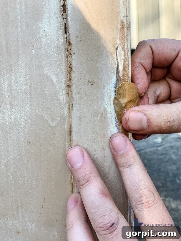 A close-up view of hands meticulously filling a damaged spot with epoxy wood filler putty, bathed in natural sunlight. The focus is on the precise application of the filler into the wood defect.