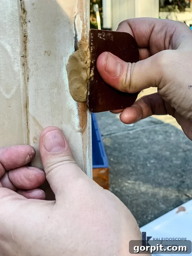 Hands meticulously use a scraper to smooth freshly applied epoxy wood filler putty onto damaged wood outdoors. The action captures the precision involved in repairing the wood surface.