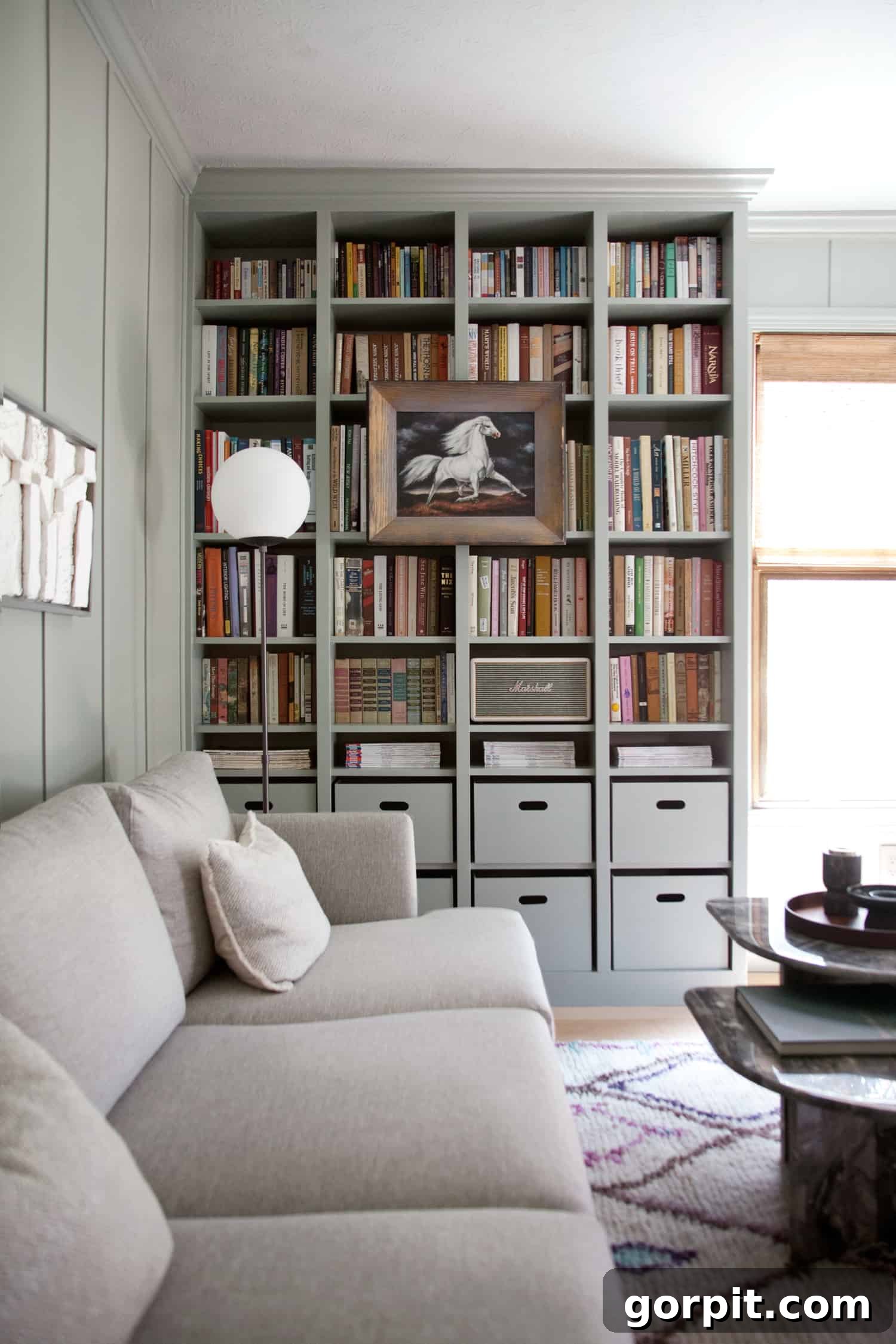 Transform Your IKEA: Bookshelf & Cabinet Built-In Hacks 9 Impressive IKEA built-in cabinets in a home library, filled with floor-to-ceiling books. The lower two shelves feature a neat row of cubby boxes, providing organized storage. A cozy gray couch rests against the connecting wall, completing the inviting reading nook.