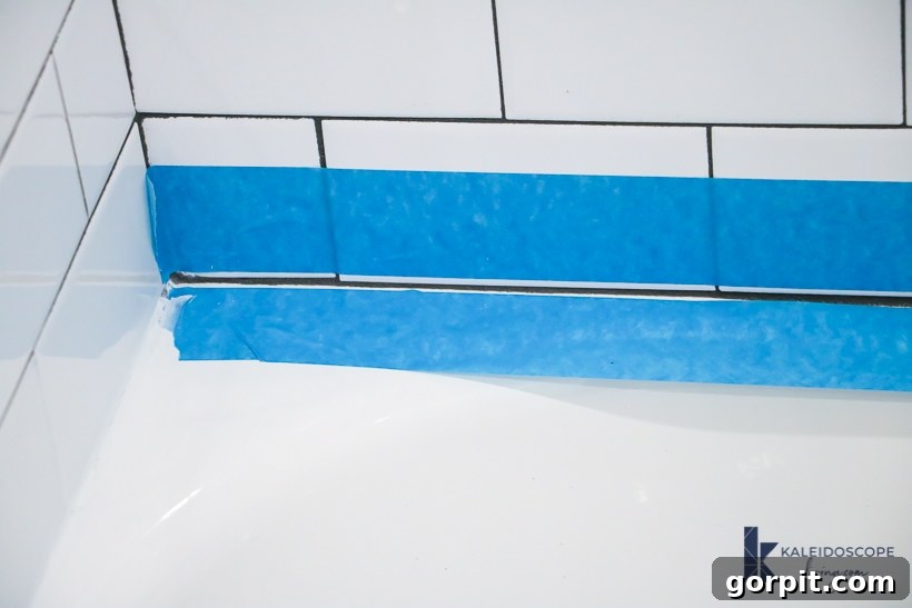 Close-up of two strips of blue painter's tape applied to white tiles, prepping a bathtub for caulking.