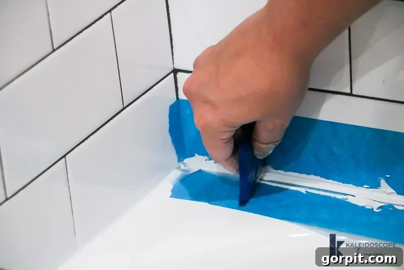 A hand uses a caulking tool to smooth caulk on a bathtub, guided by two blue strips of painter's tape.