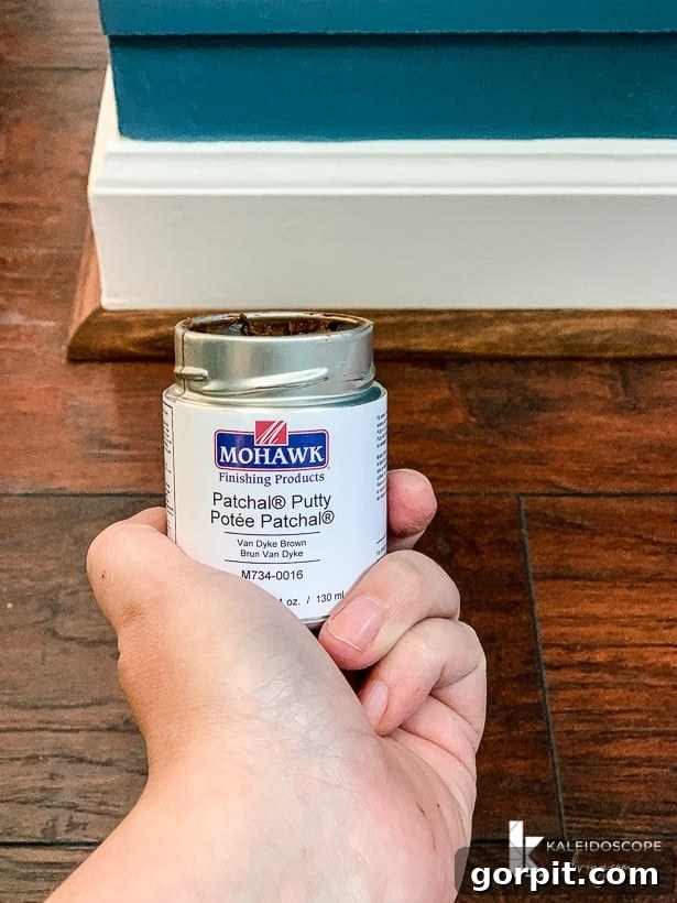 Your Guide to Perfect Quarter-Round Molding 10 A hand holds a jar of Mohawk Patchal Putty in front of quarter round molding, a white baseboard and a blue wall, preparing to fill nail holes.