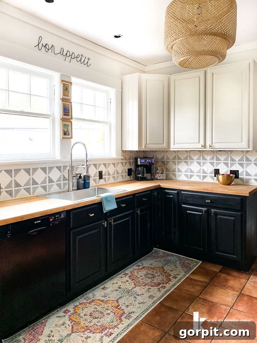 Small Budget Big Impact DIY Kitchen Transformation 13 The "after" picture of a budget-friendly kitchen makeover shows "bon appetite" on a white wall over two windows. White cabinets line the far side, above a stenciled black and white tile backsplash, with a wood counter and glossy dark green lower cabinets.