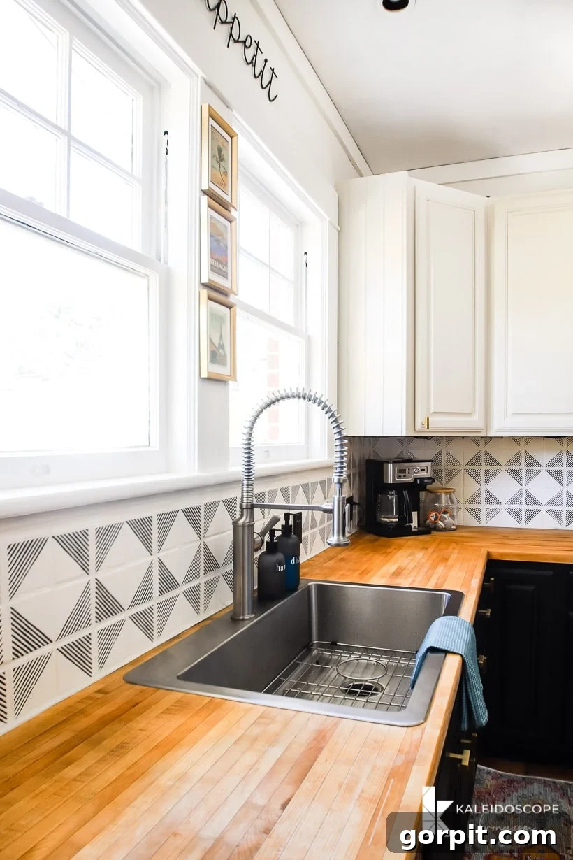 Small Budget Big Impact DIY Kitchen Transformation 16 A black and white budget-friendly kitchen makeover has geometric black and white tiles as a backsplash, a treated wood countertop, stainless steel sink, two large windows and white cabinets. A coffee machine in the corner.