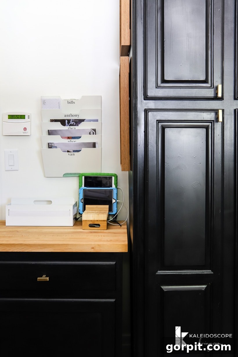 Small Budget Big Impact DIY Kitchen Transformation 17 Part of a pantry door painted black, next to a wood countertop family command center with wall-mounted file trays with family member's names and a charging station for children's tablets.