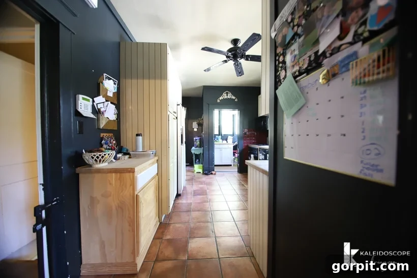Small Budget Big Impact DIY Kitchen Transformation 3 An outdated kitchen before a budget-friendly kitchen makeover removed the dark cave-like walls and trim. A family calendar hangs on the side of a refrigerator and brown tiles line the floor and hallway into the kitchen,
