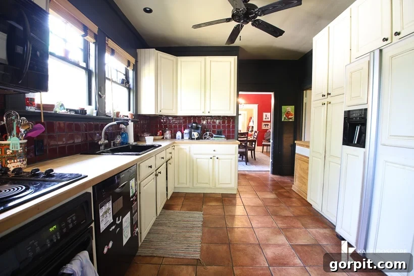 Small Budget Big Impact DIY Kitchen Transformation 4 A view into a large but narrow outdated kitchen before budget-friendly kitchen makeover. The left side holds the counter, stove, oven, sink and cabinets and the right side has a white refrigerator, pantry and upper cabinets.