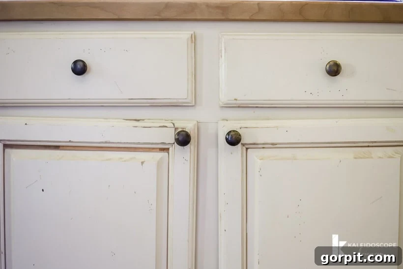 Small Budget Big Impact DIY Kitchen Transformation 6 Worn out painted kitchen cabinets with uneven hanging doors, dirt, and paint scratches. The corner text reads, "KaleidoscopeLiving.com."