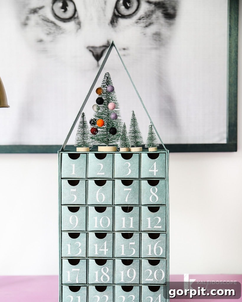 Completed metallic blue-green DIY Advent calendar with a glittery rooftop and bottlebrush trees, gracing a chic dresser.