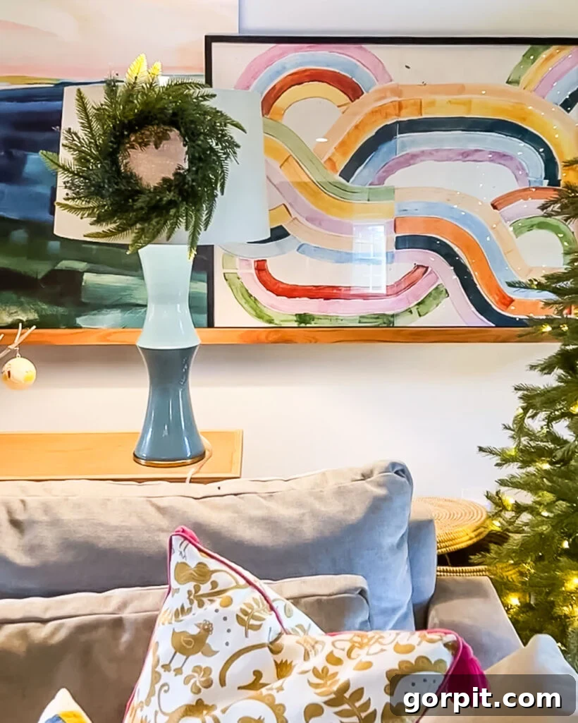 A mini wreath hanging on a lampshade in colorful family room decorated for Christmas, with the top of a gray couch and framed artwork on the wall.