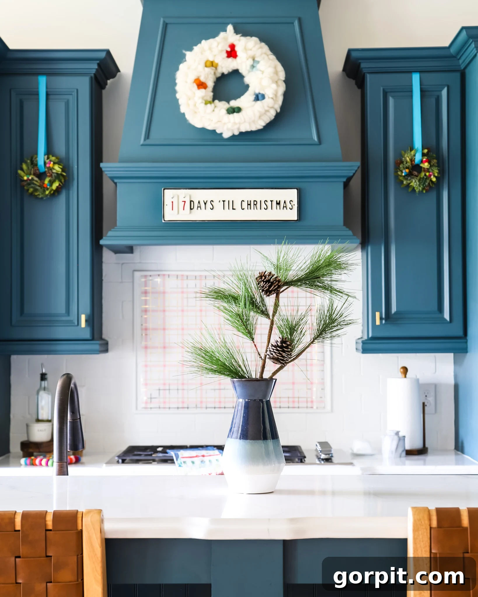 Mini wreaths hang from blue ribbons on blue cabinets and a blue exhaust hood, which are above a white-tiled backsplash and a pine tree cutting in vase in the foreground