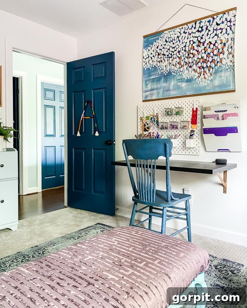 A young girl's bedroom door is painted blue to match the chair in front of a wall-mounted desk, pegboard and large art piece. 