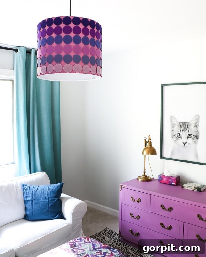 The corner of a girl's bedroom has long teal curtains, a purple shade chandelier, purple drawers and a light grey couch. 
