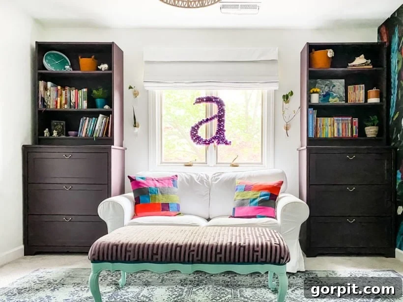 A white couch with a striped ottoman, flanked by two cabinet bookshelves and a central window with a felt ball letter "A."