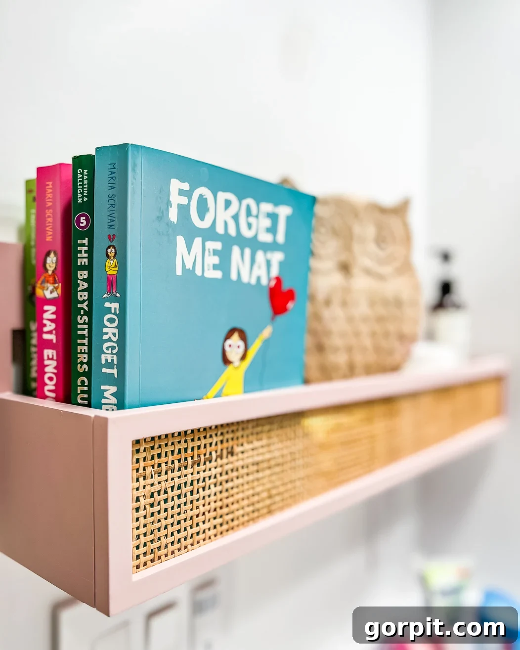 A close-up of a muted pink and caned hanging wall box full of books, and owl statue, and lotions. 