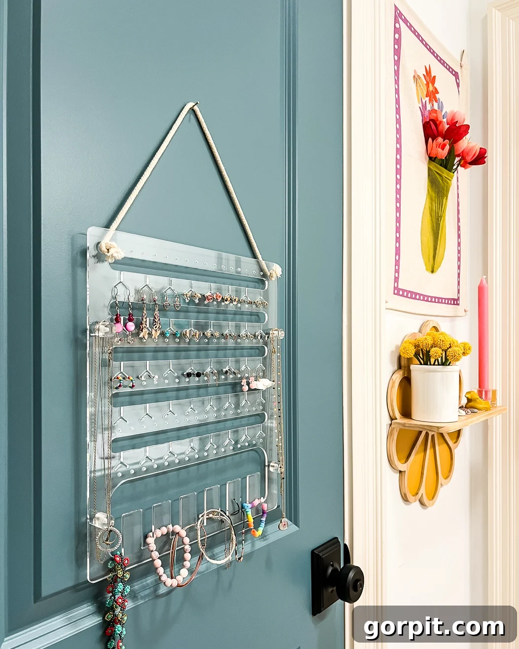 A clear acrylic hanging jewelry organizer in girl's bathroom on the back of a green-blue door with yellow flowers on the wall to the side. 