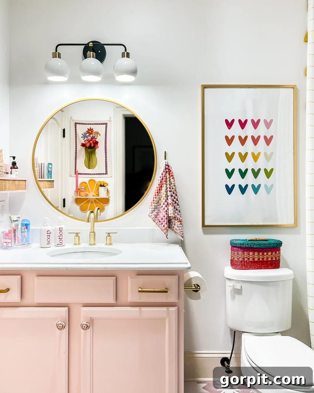 A bathroom makeover on a budget includes a round gold mirror, white walls with pink cabinets, rainbow heart artwork. toilet, and flower artwork reflected in the mirror. 