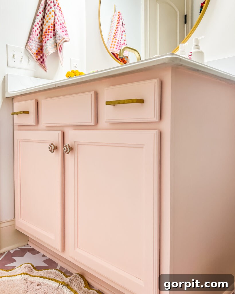 A light pink vanity in girl's bathroom, with gold hardware, a gold round mirror and white countertops. 