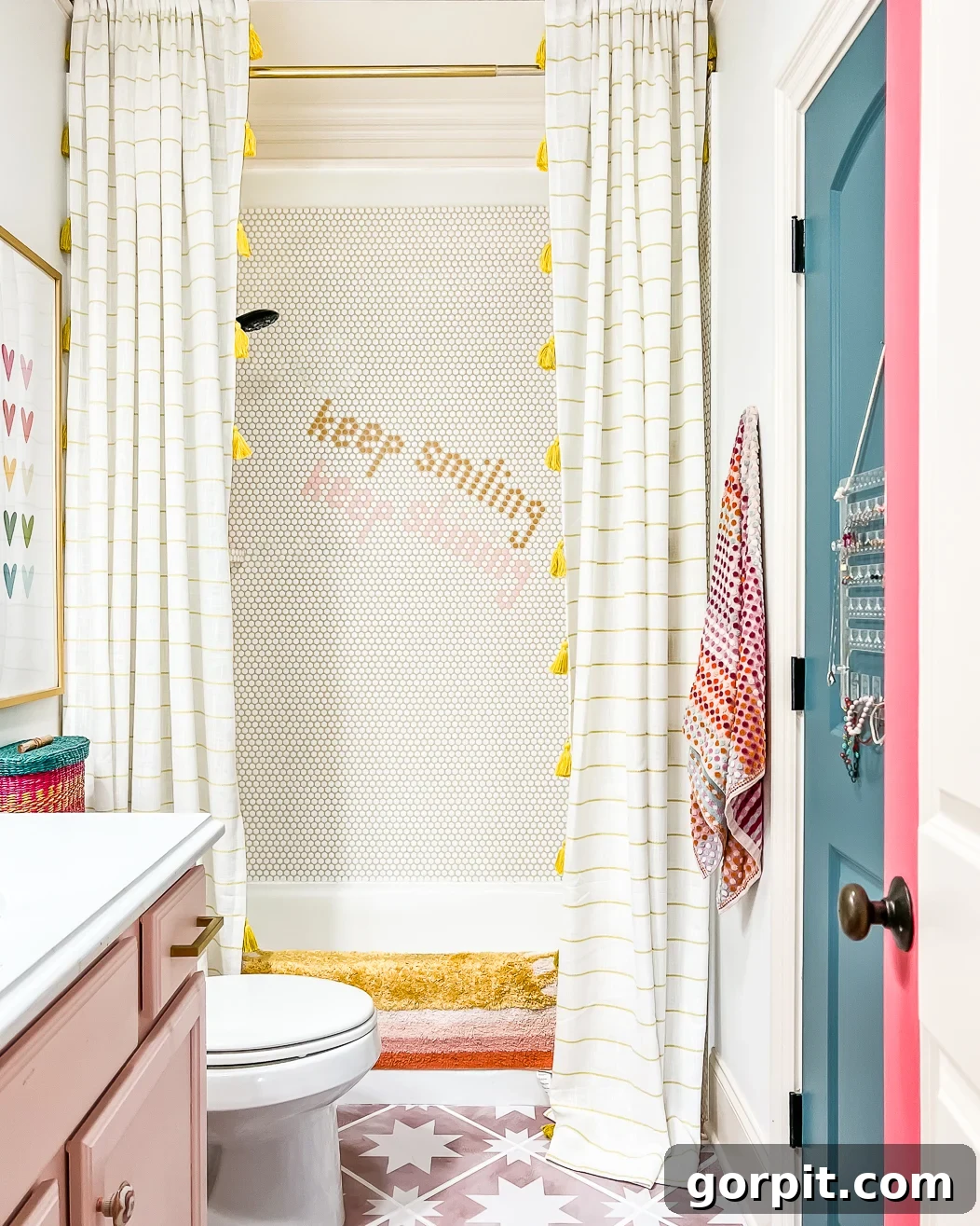 An "after" photo of a bathroom makeover on a budget features round tiles, a pink and white floor, light pink and white sink cabinet, and tub with "Keep Smiling" on the shower wall. 