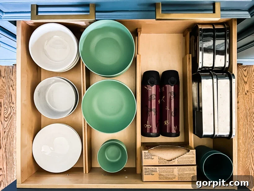 An open drawer as seen from above displays an organized kitchen drawer with stacked white and green bowls, ceramic plates, two maroon travel mugs with illustrations, stainless steel lunchboxes, paper-wrapped food items, and a green cup all separated by bamboo drawer dividers.