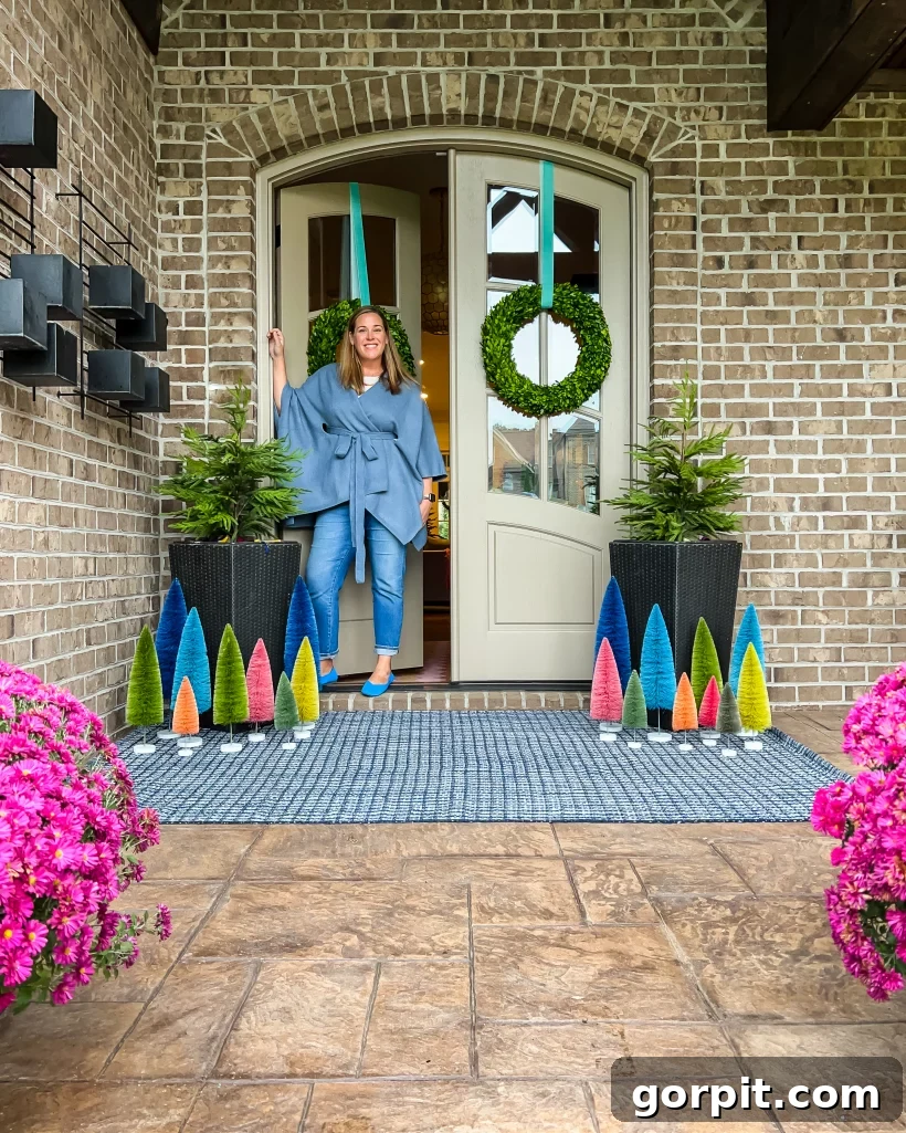 Tasha Agruso stands on her front porch among Colorful front porch Christmas decorations, including vibrant colorful bottle brush trees of various sizes, mini evergreen trees in large planters and wreaths on the front door. 