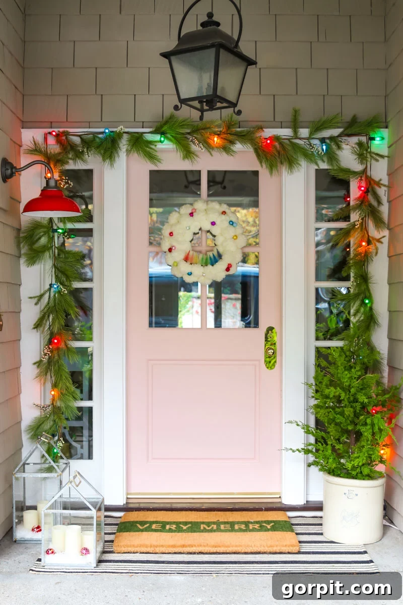 A blush-colored door in the middle of large brick walls has a sparse garland with multicolored lights framing the door, and other front porch Christmas decorations including a mini evergreen in a white plants, and candles in glass cases. These front porch Christmas decorations were done by Modern Glam. 