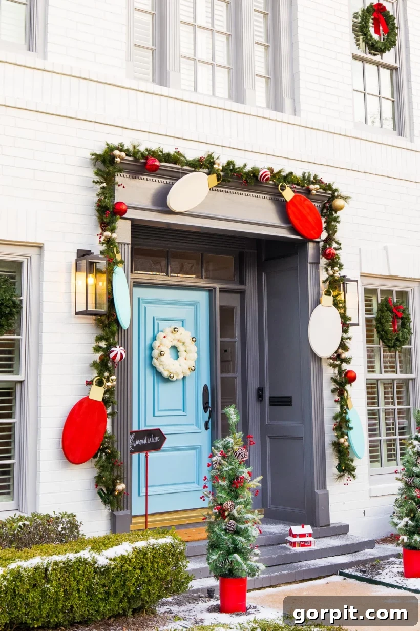 Front porch Christmas decorations by Sugar and Cloth include oversized light bulb cutouts in red, white, and teal hanging from garland that frames the entry. A bright turquoise front door features a fluffy white wreath. Two mini-Christmas trees in red pots are on either side of the steps.