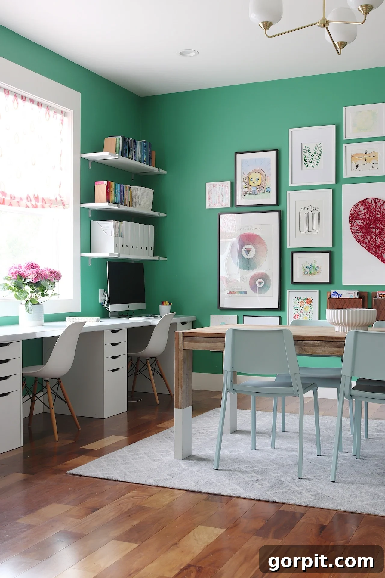 A green room has white desks for homework against the windows and a craft table in the middle of the room/ Th walls are covered in framed pictures and art, which is just one of many playroom ideas. 